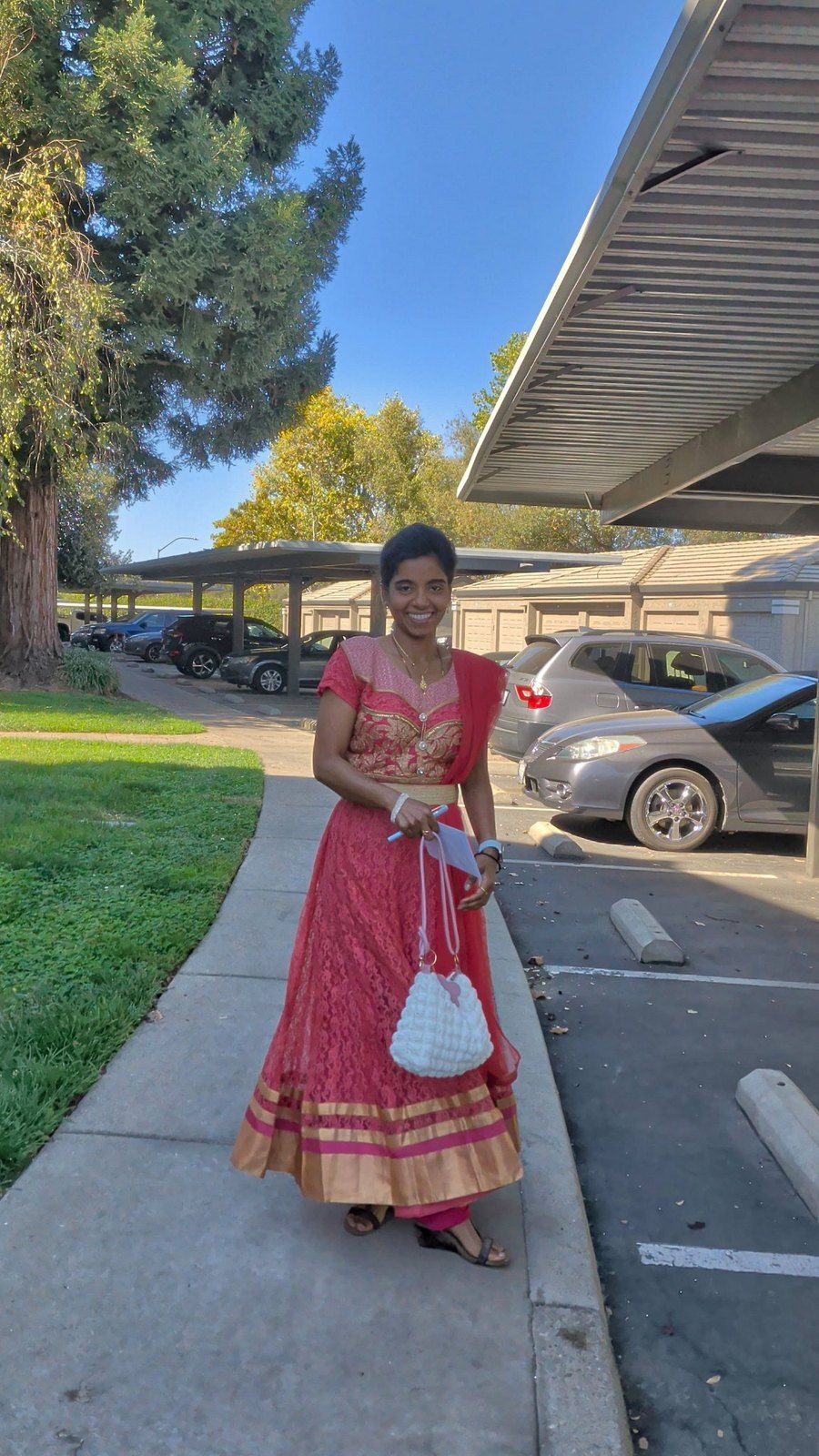 Nithya today, smiling in the same coral-pink dress outdoors on a sunny day